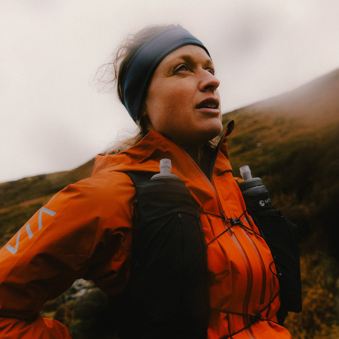 A woman in a mountain setting wearing an orange running jacket, a black headband, and a hydration pack with water bottles.